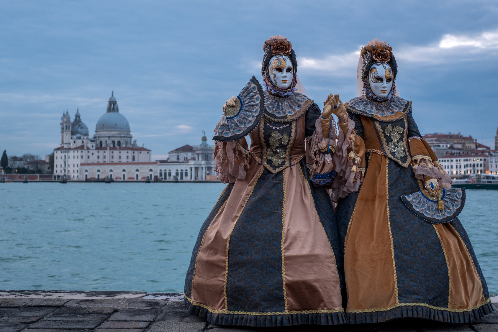 Two women in traditional costumes and masks, standing in front of the Grand Canal during the Venice Carnival. Basilica di Santa Maria della Salute in the background